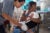 A female health care worker wearing blue scrubs inserts a vaccine syringe into a toddler’s leg.The toddler is being held by her mother who is seated in a clinic room.