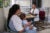 A female health care worker wearing a white coat sits at a desk in a red chair and talks with a mother who holds an infant in her lap in northeastern India.