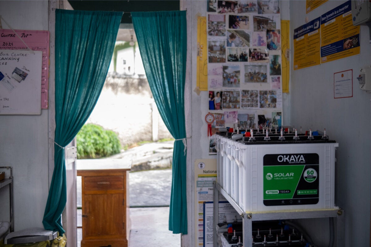 A row of batteries that store solar power sit on a grey metal shelf in a room in a health clinic in northeastern India. The room has a green curtain on the doorway and various community photos and signs on the walls.